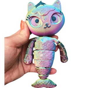 May include: A colorful, articulated cat figurine with a mermaid tail. The cat's head and body are a blend of iridescent colors, including blue, purple, and yellow. The figurine is held in a hand, showcasing its flexible joints and unique design.