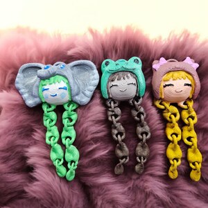 May include: Three whimsical character pendants, each with a unique animal hat design: an elephant, a frog, and a bear. The pendants feature chain-like strands in green, gray, and yellow, respectively. The characters have smiling faces.