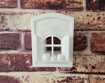 May include: A white miniature window with an arched top, shutters, and a small shelf. The window has a cross-shaped pane and three small, round decorative objects on the shelf. The background is a brick wall.