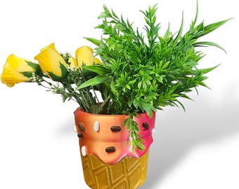 Könnte beinhalten: Ein dekorativer Pflanztopf in Form einer Eistüte, gefüllt mit gelben Rosen und grünem Laub. Die Tüte ist goldfarben, das Eis ist orange und rosa und mit weißen und braunen Streuseln verziert. Ein einzigartiges Blumenarrangement.