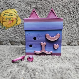 May include: A small, purple and pink cat-shaped box with a heart, a crescent moon, and a nose on the front. The box is made of 3D printed plastic and has a textured surface.