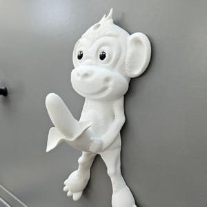 May include: White 3D printed monkey figurine holding a banana. The monkey has a playful expression and is holding the banana in its right hand.