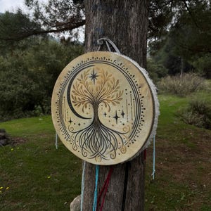 Peut inclure: Un tambour rond beige orn&eacute; d'un motif d'arbre de vie, d'un croissant de lune et d'&eacute;toiles. Le tambour est suspendu &agrave; un tronc d'arbre avec des cordes color&eacute;es. Il a une bordure en fourrure blanche.