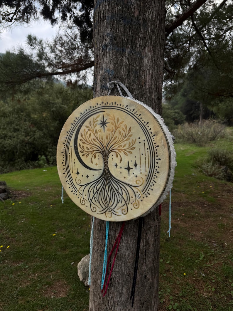 Peut inclure: Un tambour rond beige orn&eacute; d'un motif d'arbre de vie, d'un croissant de lune et d'&eacute;toiles. Le tambour est suspendu &agrave; un tronc d'arbre avec des cordes color&eacute;es. Il a une bordure en fourrure blanche.