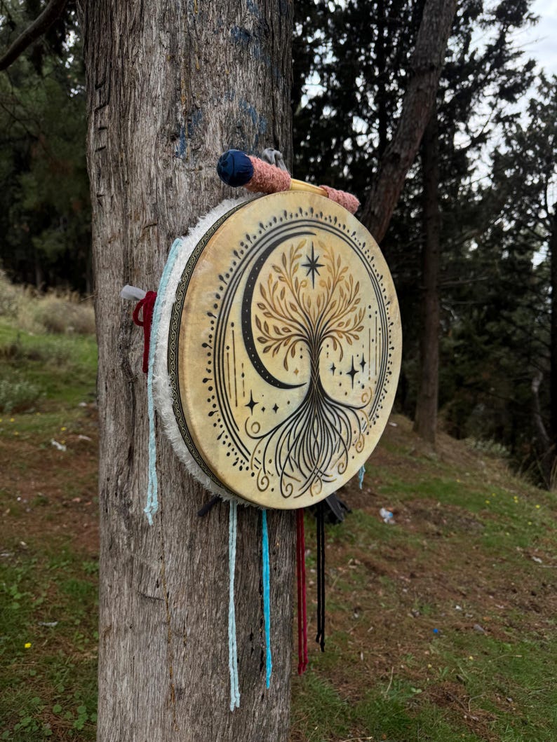 Peut inclure: Un tambourin rond beige orn&eacute; d'un motif d'arbre de vie, d'une lune et d'&eacute;toiles. Le tambour est suspendu &agrave; un tronc d'arbre. Une baguette avec une t&ecirc;te bleue et rose est pos&eacute;e dessus. Des cordes color&eacute;es pendent du c&ocirc;t&eacute;.