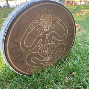 Op de afbeelding: Een ronde houten trommel met een gedetailleerd ontwerp van een zon-figuur. De trommel heeft een bruin oppervlak met een lichtgekleurde illustratie. De rand van de trommel is wit en zacht. De trommel ligt op groen gras.
