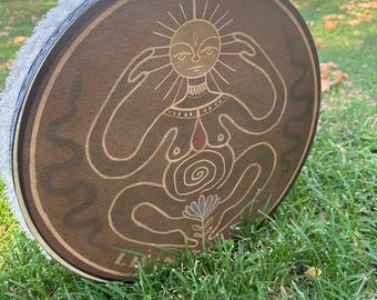 May include: A round, wooden drum with a detailed sun-faced figure design. The drum's face is brown with a light-colored illustration. The drum's rim is white and fluffy. The drum is resting on green grass.
