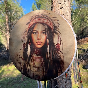 Puede incluir: Un tambor redondo de cuero marr&oacute;n con una franja blanca alrededor del borde. El tambor est&aacute; decorado con una imagen colorida de una mujer nativa americana con un tocado.