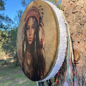 Puede incluir: Un tambor de cuero marr&oacute;n con ribete de piel blanca y borlas de colores. El tambor presenta una imagen impresa de una mujer nativa americana con tocado.
