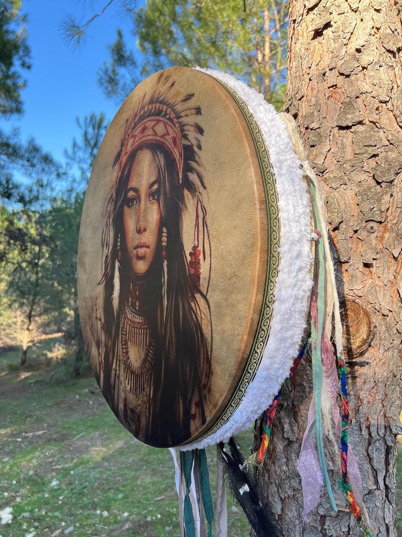 Puede incluir: Un tambor de cuero marr&oacute;n con ribete de piel blanca y borlas de colores. El tambor presenta una imagen impresa de una mujer nativa americana con tocado.