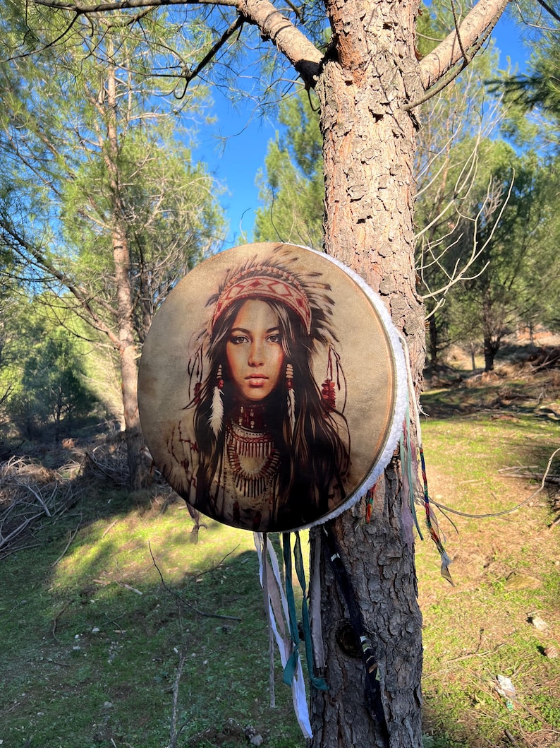 Puede incluir: Un tambor redondo de cuero marr&oacute;n con una franja blanca que cuelga de la parte inferior. El tambor est&aacute; decorado con una imagen colorida de una mujer nativa americana con un tocado. El tambor est&aacute; colgado de un tronco de &aacute;rbol en un entorno boscoso.