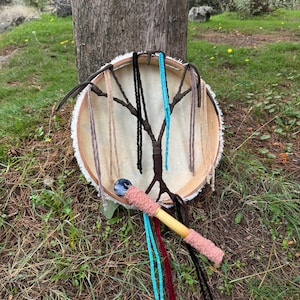 Peut inclure: Un tambour artisanal avec une peau de couleur claire tendue sur un cadre en bois. Le tambour pr&eacute;sente un motif en forme d'arbre avec des cordons suspendus de diff&eacute;rentes couleurs. Un b&acirc;ton de tambour en bois avec une poign&eacute;e rose et des rubans color&eacute;s est &agrave; proximit&eacute;.