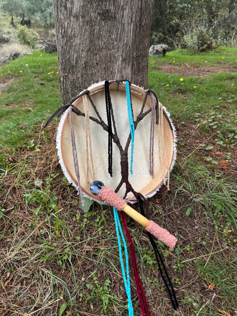 Peut inclure: Un tambour artisanal avec une peau de couleur claire tendue sur un cadre en bois. Le tambour pr&eacute;sente un motif en forme d'arbre avec des cordons suspendus de diff&eacute;rentes couleurs. Un b&acirc;ton de tambour en bois avec une poign&eacute;e rose et des rubans color&eacute;s est &agrave; proximit&eacute;.