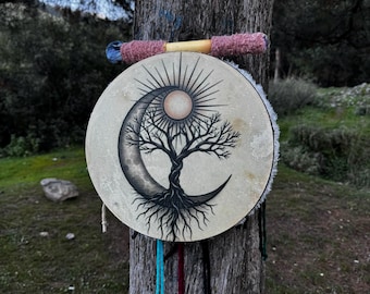 May include: A round drum with a tree of life design, featuring a crescent moon and sun. The drum is beige with a black illustration and is adorned with colorful hanging cords. A pink and blue mallet rests on top.