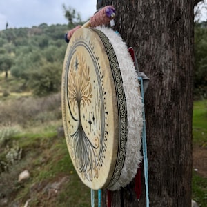 Peut inclure: Un tambour rond peint &agrave; la main avec un motif d'arbre de vie. La peau du tambour est beige avec une illustration d'arbre noir et or. Le bord est orn&eacute; d'un motif noir et blanc et de franges blanches. Des cordons color&eacute;s et une baguette sont attach&eacute;s.