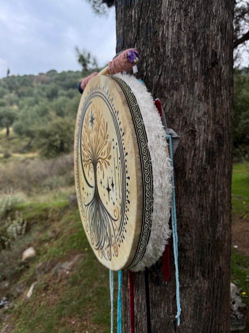 Peut inclure: Un tambour rond peint &agrave; la main avec un motif d'arbre de vie. La peau du tambour est beige avec une illustration d'arbre noir et or. Le bord est orn&eacute; d'un motif noir et blanc et de franges blanches. Des cordons color&eacute;s et une baguette sont attach&eacute;s.