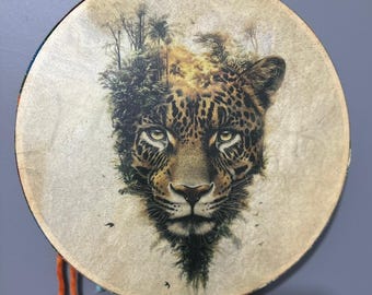 May include: A round drum with a detailed illustration of a jaguar's face and a jungle scene. The jaguar has a spotted coat and intense eyes. The drum's rim is decorated with colorful fabric and hanging strands.