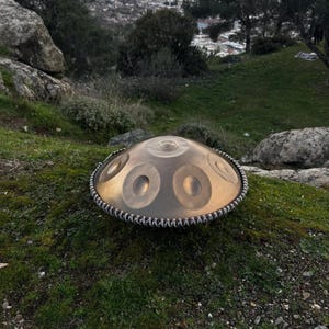 May include: A bronze-colored handpan drum with multiple circular sound holes, resting on a grassy hillside. The instrument has a decorative border and is set against a backdrop of a distant cityscape and natural landscape.