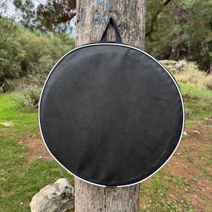 Peut inclure: Un sac rond noir avec une bordure blanche et une poign&eacute;e noire est suspendu &agrave; un tronc d'arbre. Le sac a un design simple et fonctionnel. L'arri&egrave;re-plan pr&eacute;sente une sc&egrave;ne ext&eacute;rieure naturelle avec des arbres et de la verdure.