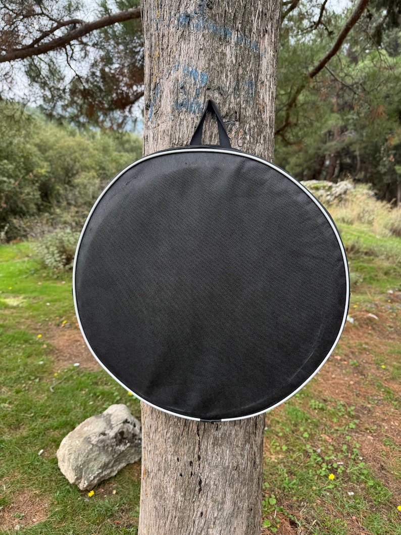 Peut inclure: Un sac rond noir avec une bordure blanche et une poign&eacute;e noire est suspendu &agrave; un tronc d'arbre. Le sac a un design simple et fonctionnel. L'arri&egrave;re-plan pr&eacute;sente une sc&egrave;ne ext&eacute;rieure naturelle avec des arbres et de la verdure.