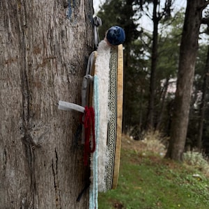 Peut inclure: Un tambour traditionnel avec un cadre en bois et une bordure &agrave; motifs est attach&eacute; &agrave; un arbre. Le tambour a une couverture en tissu bleu et blanc et est orn&eacute; de pompons rouges et bleus. L'arri&egrave;re-plan pr&eacute;sente un cadre forestier.