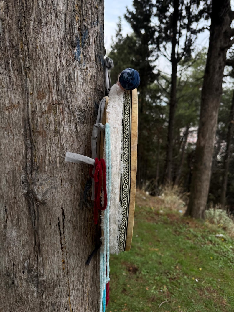 Peut inclure: Un tambour traditionnel avec un cadre en bois et une bordure &agrave; motifs est attach&eacute; &agrave; un arbre. Le tambour a une couverture en tissu bleu et blanc et est orn&eacute; de pompons rouges et bleus. L'arri&egrave;re-plan pr&eacute;sente un cadre forestier.