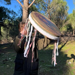 Puede incluir: Una persona sostiene un tambor de marco grande, redondo y de madera con un borde de tela blanca y borlas de tela de colores que cuelgan de la parte inferior. El tambor tiene un dise&ntilde;o pintado en la parte delantera.