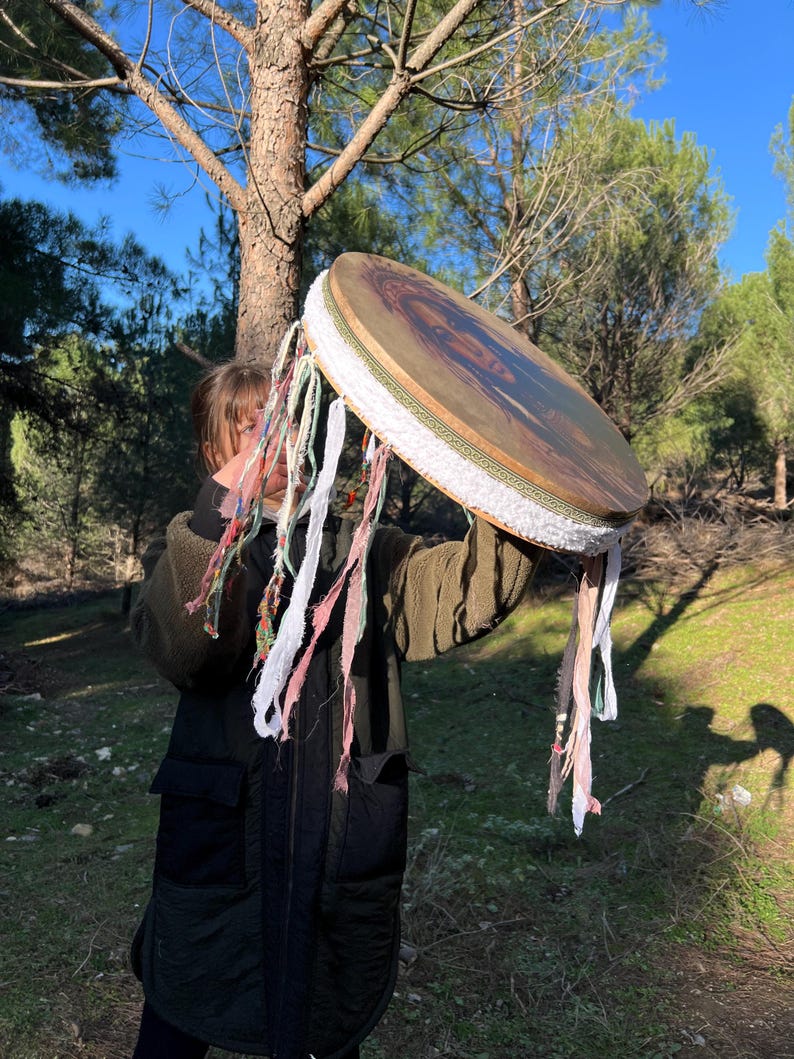 Puede incluir: Una persona sostiene un tambor de marco grande, redondo y de madera con un borde de tela blanca y borlas de tela de colores que cuelgan de la parte inferior. El tambor tiene un dise&ntilde;o pintado en la parte delantera.