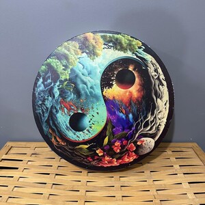 Op de afbeelding: Een ronde, houten, decoratieve wandhanger met een kleurrijk yin yang-ontwerp. Het ontwerp heeft een blauwe en groene kant met een zwart-witte maan en een rode en oranje kant met een zwart-witte zon.