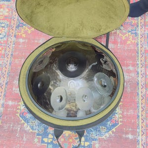 May include: A silver handpan drum with multiple circular sound fields, resting inside an open black case with a green interior. The handpan has a central dome and is surrounded by several circular indentations. The case has a black handle.