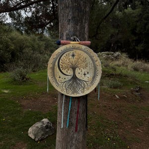 Peut inclure: Un tambour rond beige orn&eacute; d'un motif d'arbre de vie, d'un croissant de lune et d'&eacute;toiles est suspendu &agrave; un tronc d'arbre. Le tambour pr&eacute;sente une illustration d&eacute;taill&eacute;e. Une baguette avec des accents roses et bleus repose au-dessus.