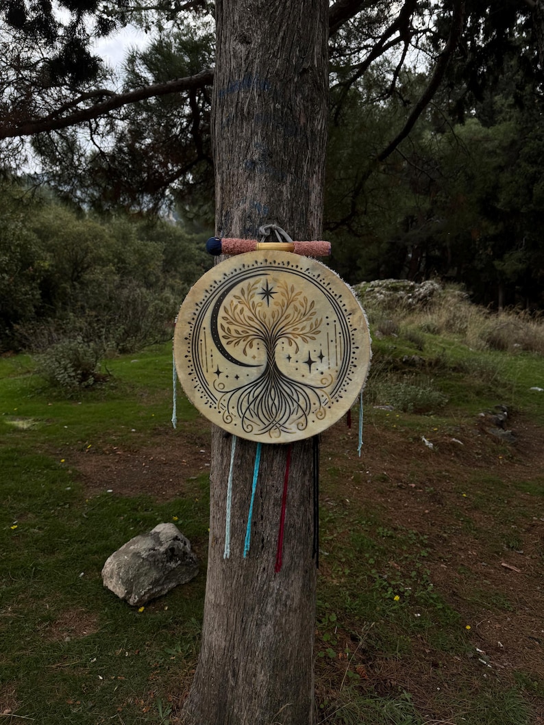 Peut inclure: Un tambour rond beige orn&eacute; d'un motif d'arbre de vie, d'un croissant de lune et d'&eacute;toiles est suspendu &agrave; un tronc d'arbre. Le tambour pr&eacute;sente une illustration d&eacute;taill&eacute;e. Une baguette avec des accents roses et bleus repose au-dessus.
