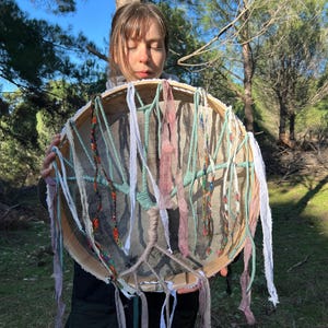 Puede incluir: Una mujer sostiene un tambor grande y redondo con una cabeza de cuero natural. El tambor est&aacute; decorado con cintas de tela de colores y cuentas. El tambor est&aacute; rodeado de &aacute;rboles y hierba verde.