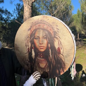 Puede incluir: Un tambor redondo con una franja blanca alrededor del borde. El tambor est&aacute; decorado con una imagen colorida de una mujer nativa americana con un tocado. El tambor se toca con un palo de madera con una cabeza rayada blanca y negra.