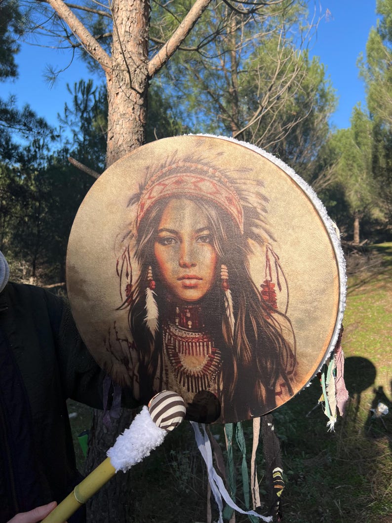 Puede incluir: Un tambor redondo con una franja blanca alrededor del borde. El tambor est&aacute; decorado con una imagen colorida de una mujer nativa americana con un tocado. El tambor se toca con un palo de madera con una cabeza rayada blanca y negra.