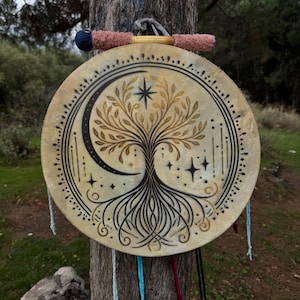 Peut inclure: Un tambourin rond beige avec un motif peint &agrave; la main d'arbre de vie, un croissant de lune et des &eacute;toiles. Le tambourin est suspendu &agrave; un tronc d'arbre par une poign&eacute;e rose et bleue. Le tambourin est d&eacute;cor&eacute; d'accents noirs et dor&eacute;s.