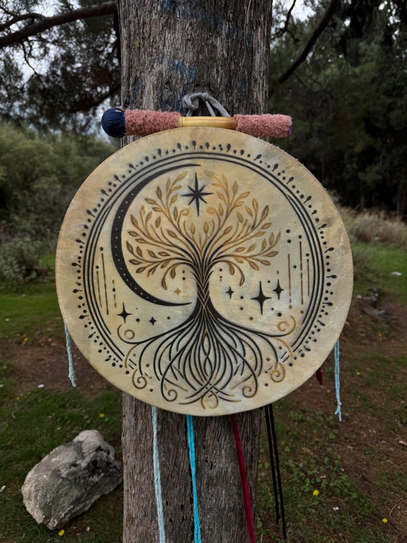Peut inclure: Un tambourin rond beige avec un motif peint &agrave; la main d'arbre de vie, un croissant de lune et des &eacute;toiles. Le tambourin est suspendu &agrave; un tronc d'arbre par une poign&eacute;e rose et bleue. Le tambourin est d&eacute;cor&eacute; d'accents noirs et dor&eacute;s.