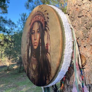 Puede incluir: Un tambor redondo de cuero marr&oacute;n con un ribete de piel blanca y cintas de colores que cuelgan de la parte inferior. El tambor presenta una imagen impresa de una mujer nativa americana con un tocado.