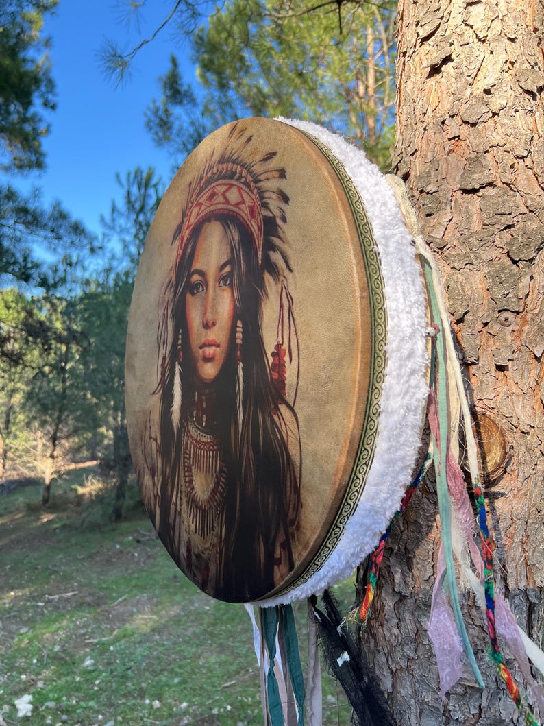 Puede incluir: Un tambor redondo de cuero marr&oacute;n con un ribete de piel blanca y cintas de colores que cuelgan de la parte inferior. El tambor presenta una imagen impresa de una mujer nativa americana con un tocado.