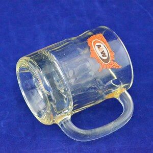 May include: A clear glass mug with a handle. The mug has a brown and orange A&W logo on it, with the letters "A&W" inside a stylized outline of the United States.