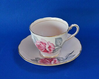 May include: A vintage teacup and saucer set. The white porcelain cup and saucer feature a pink rose design with gray leaves and gold trim. The cup has a curved handle and sits on a matching saucer, both against a blue background.