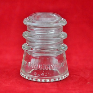 May include: A clear glass insulator with three tiers and a Hemingray 10 0-4 inscription. The insulator is on a red background.