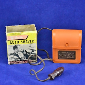 Op de afbeelding: Een vintage oranje elektrische reis scheerapparaat in de originele doos. Het scheerapparaat heeft een snoer en een sigarettenaansteker stekker. De doos heeft een afbeelding van een man die zich scheert in een auto.
