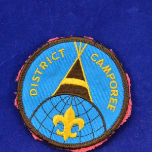May include: A circular patch with the words "DISTRICT CAMPOREE" in yellow lettering on a blue background. The patch features a brown and yellow teepee design, a gold fleur-de-lis, and a blue globe design.