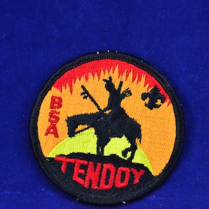Op de afbeelding: Een geborduurde patch in zwart en rood met een silhouet van een cowboy die op een paard rijdt tegen een gele en oranje zonsondergang. De tekst "BOGA TENDOY" staat in rood onderaan de patch.