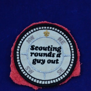 May include: A white embroidered patch with a compass design and the text "Scouting rounds a guy out". The patch is surrounded by a black border and a red fabric backing.
