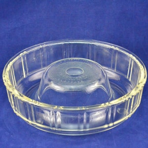 Puede incluir: Molde para bizcocho bundt de vidrio transparente con un tubo central y un diseño exterior acanalado. El molde se encuentra sobre una superficie azul. Está diseñado para hornear pasteles y otros postres.