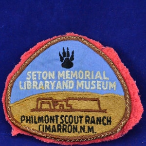 Może przedstawiać: Naszywka haftowana w kolorze brązowym i beżowym na niebieskim tle. Naszywka przedstawia czarną odbitkę łapy, tekst "SETON MEMORIAL LIBRARY AND MUSEUM" oraz tekst "PHILMONT SCOUT RANCH CIMARRON, N.M."
