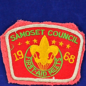 Könnte beinhalten: Ein gestickter Aufnäher in Rot, Weiß und Grün mit dem Text "Samoset Council" und "First Aid Meets" in einem Kreis um eine gelbe Lilie. Die Zahlen "19" und "68" befinden sich auf beiden Seiten des Kreises.