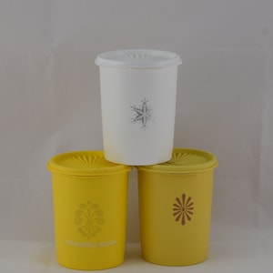 Puede incluir: Tres recipientes de almacenamiento de alimentos vintage. Un recipiente blanco con un diseño de copo de nieve y dos recipientes amarillos con diseños decorativos. Un recipiente amarillo tiene la inscripción "powdered sugar".